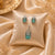 Emerald Élan American Diamond pendant set with emerald green rectangular stones, matching earrings, and sparkling halo detailing in gold polish.